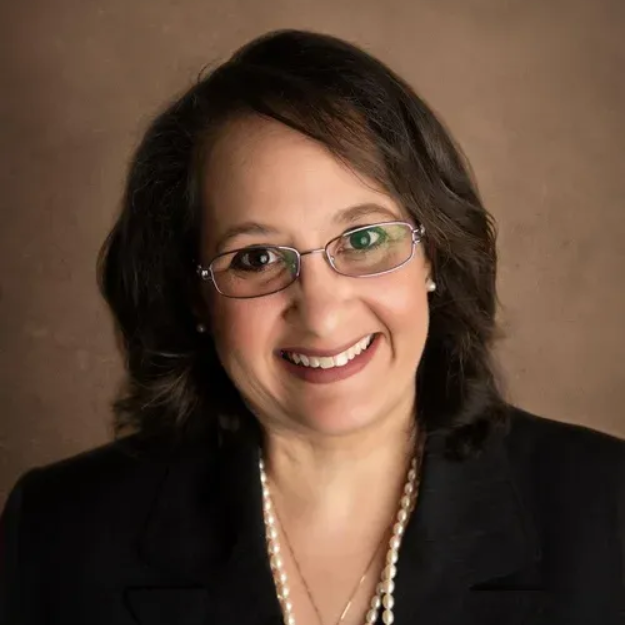 A person wearing a blue checkered blazer and light pink shirt, smiling against a soft-focus green background.