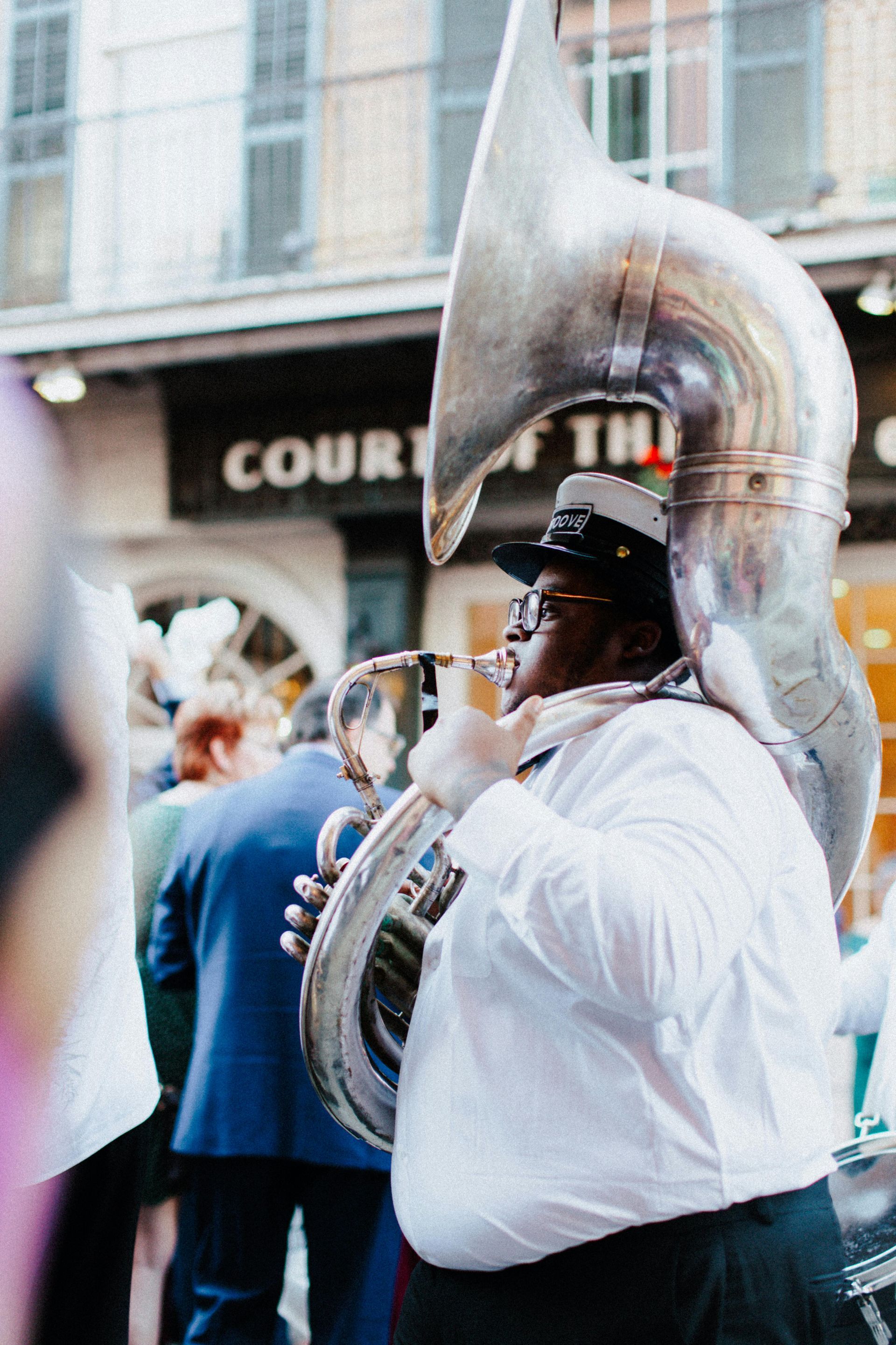 A musician in a white shirt plays a large silver sousaphone during an outdoor street parade.