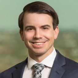 A person with a dark beard and styled hair, wearing a green button-down shirt, smiles against a plain light background.