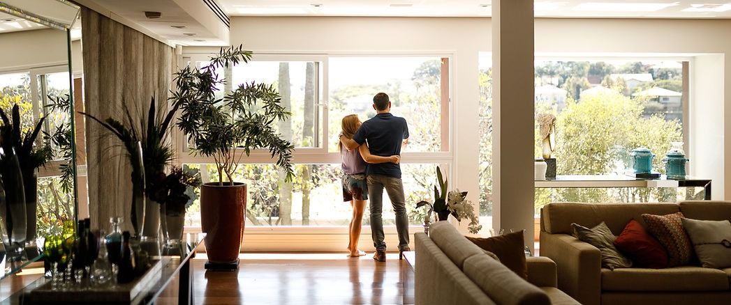 Couple standing together in a sunlit living room, looking out large windows toward a garden.