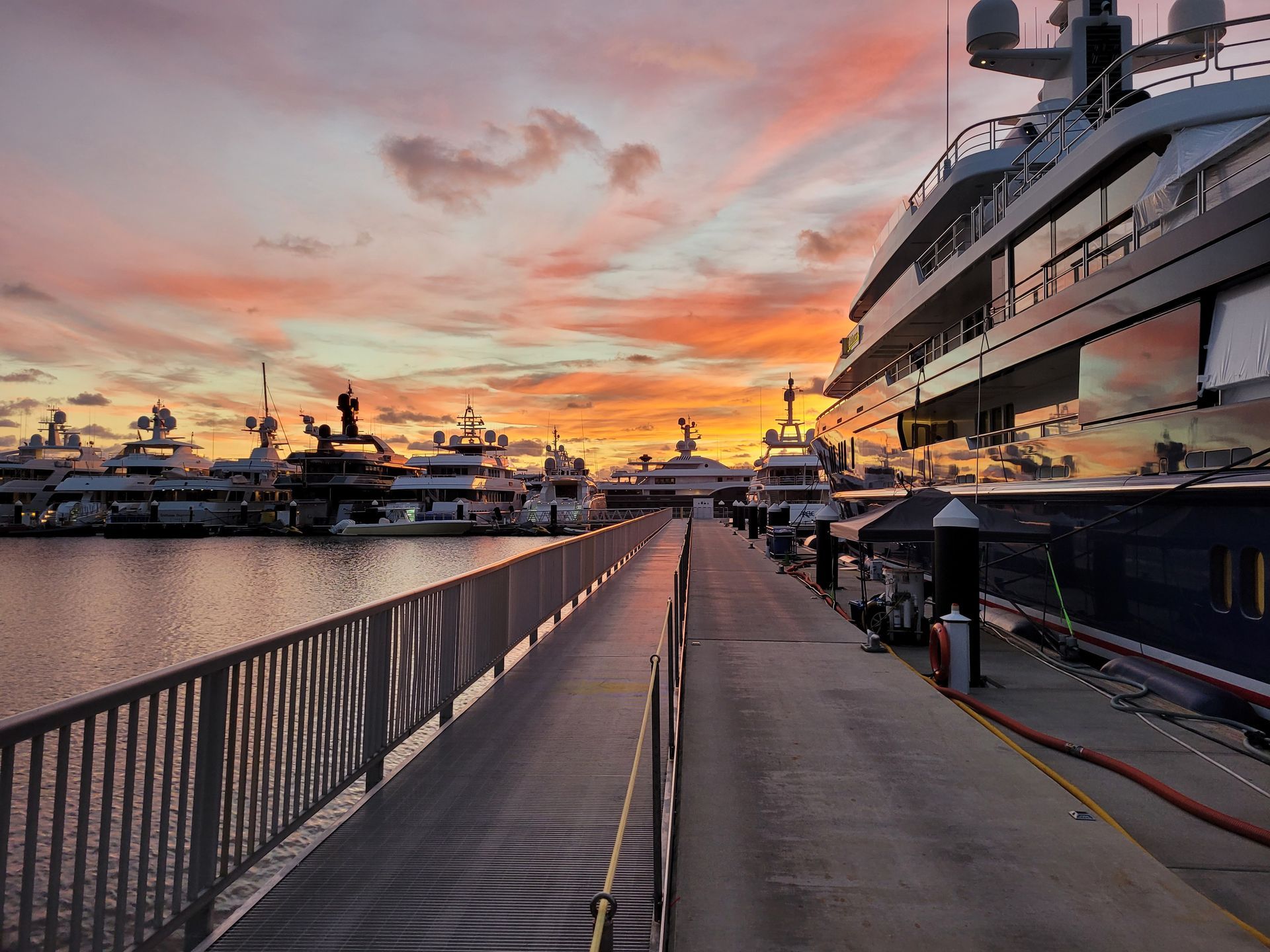 A marina with large yachts docked at a concrete pier during a vibrant, colorful sunset with orange and purple clouds.