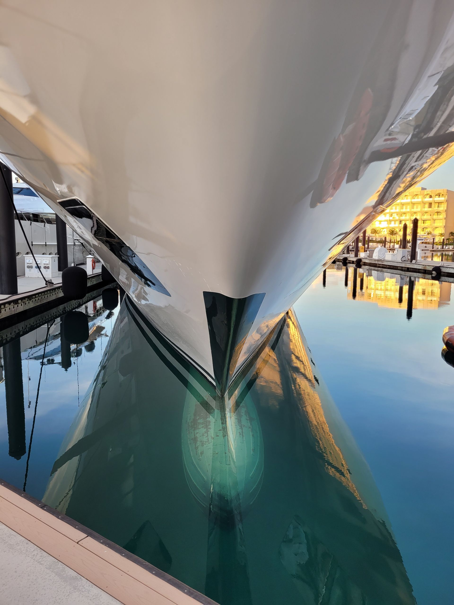 The bow of a white yacht reflects on the calm, blue water of a marina at sunset.