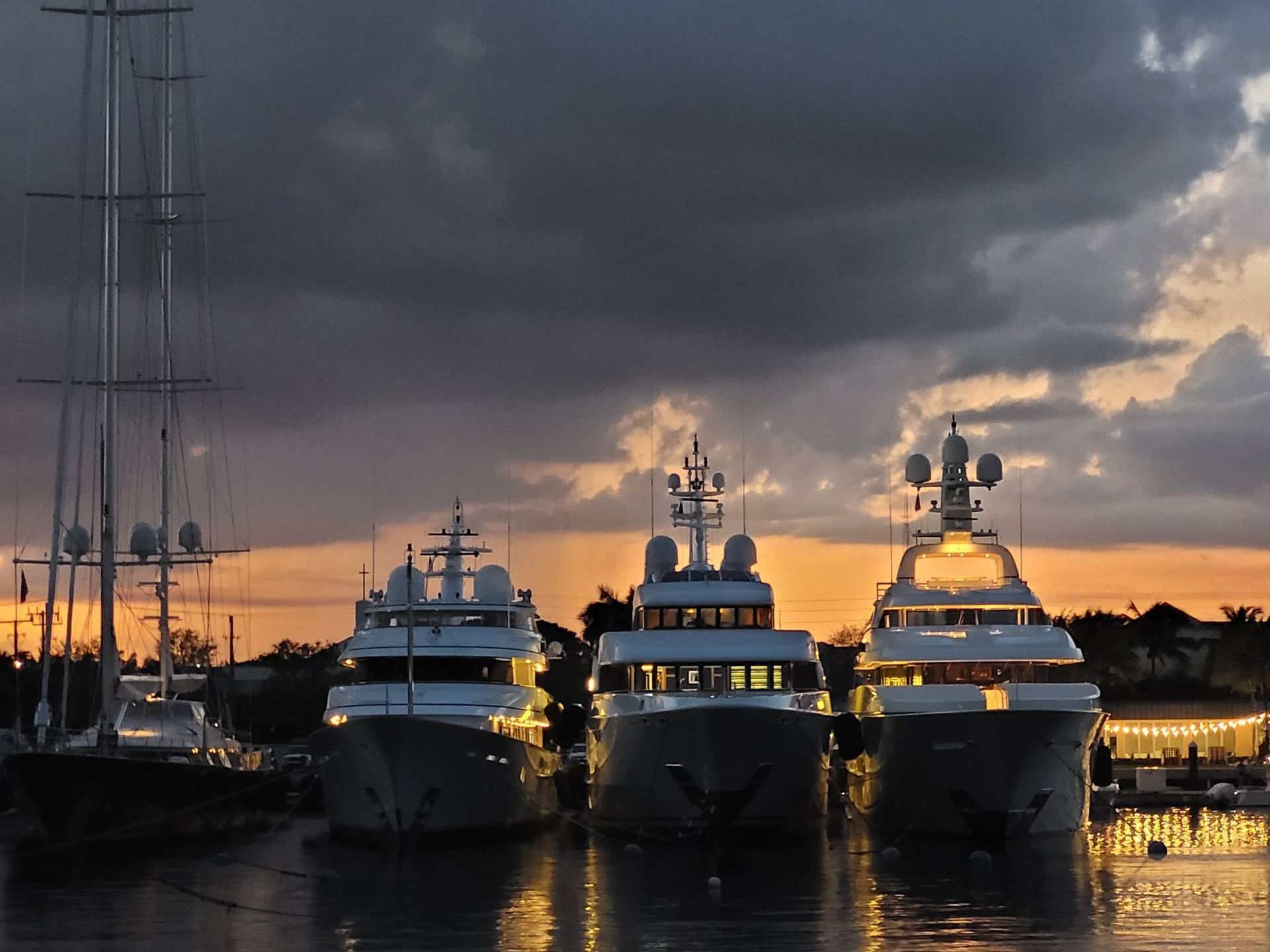 Three large yachts moored in a harbor at sunset, with dark clouds above and soft orange light on the water.