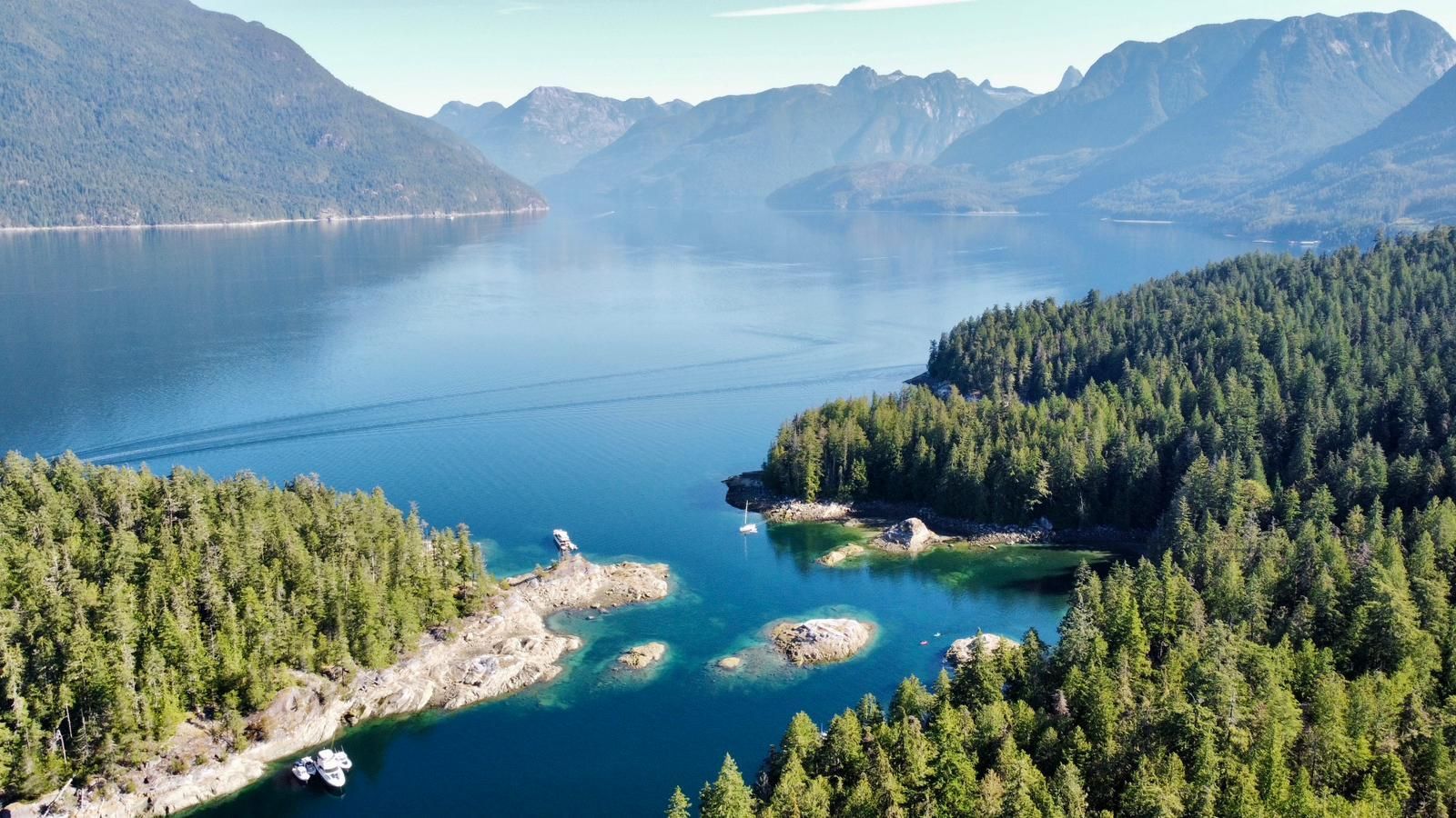 Aerial view of a calm blue inlet surrounded by dense green forests, rugged rocky outcrops, and distant mountains.