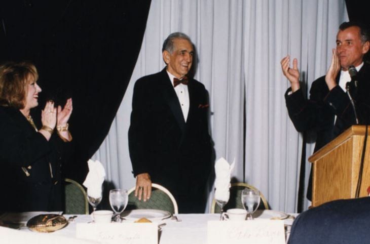A man in a tuxedo stands at a podium while a woman applauds
