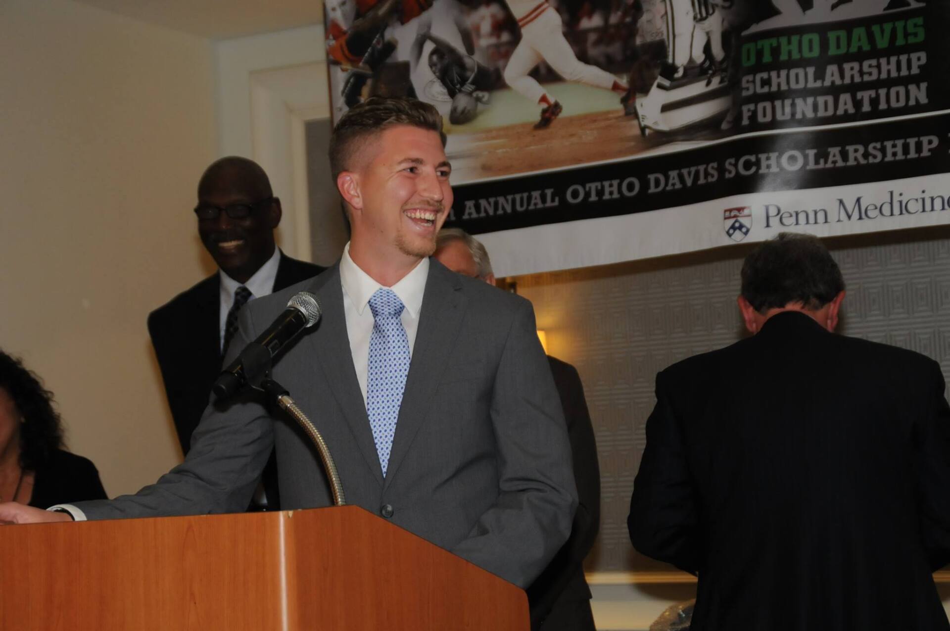 A man in a suit and tie stands at a podium speaking into a microphone