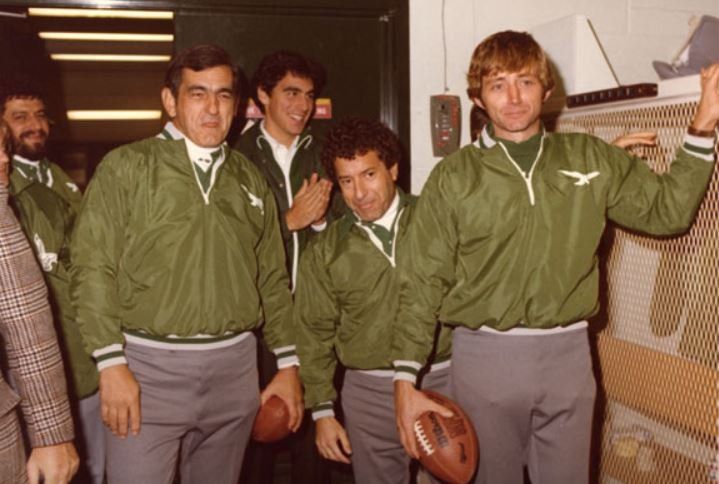 A group of men wearing green jackets and grey pants are holding footballs