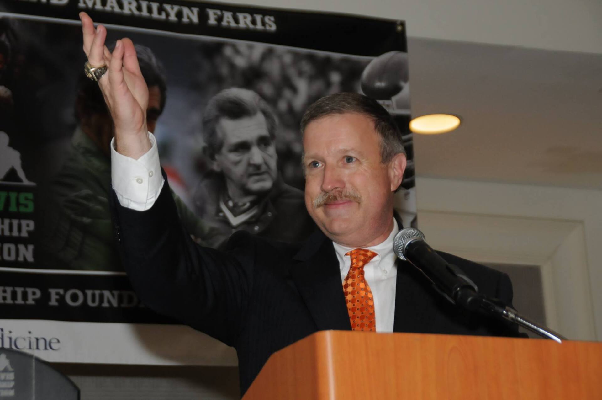 A man stands at a podium with a marilyn faris poster behind him