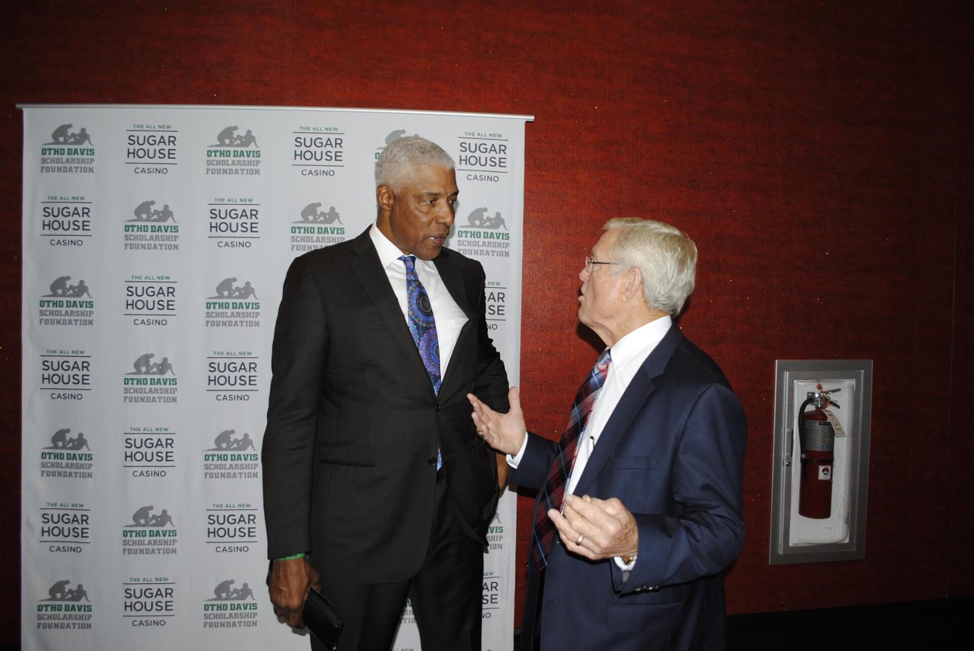 Two men in suits and ties are talking in front of a wall with a fire extinguisher on it