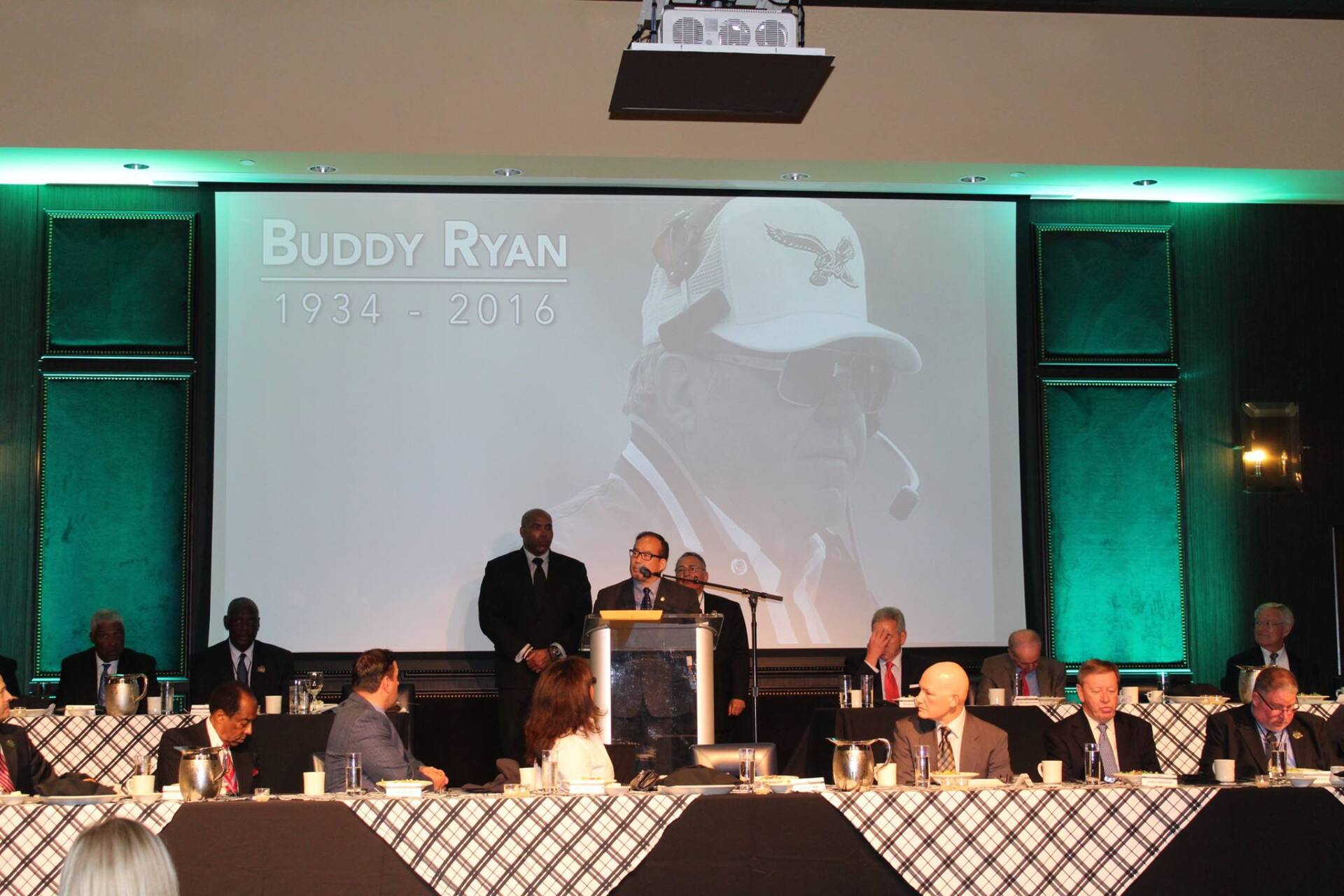 A man stands at a podium in front of a large screen that says buddy ryan