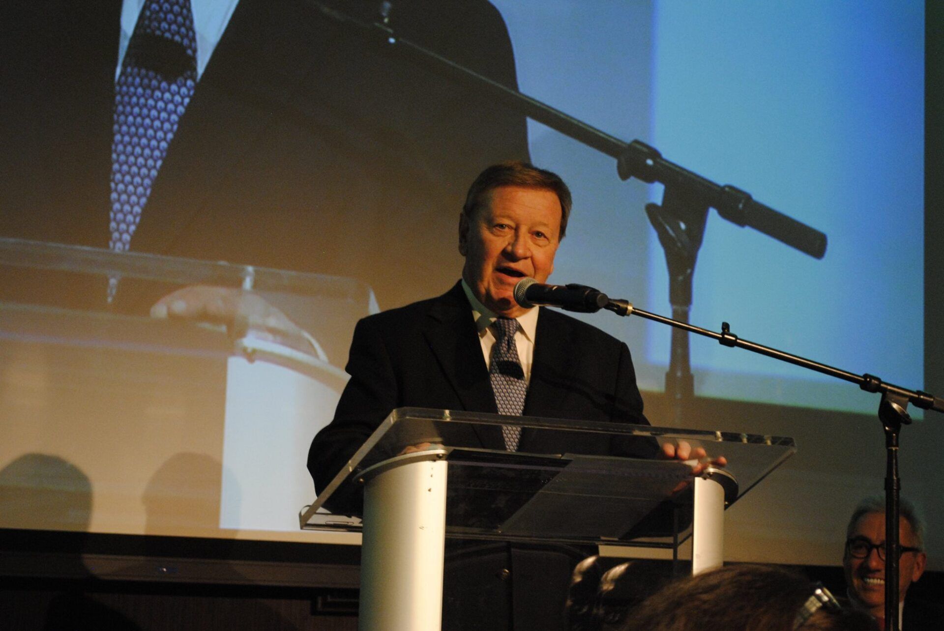 A man in a suit and tie is standing at a podium speaking into a microphone