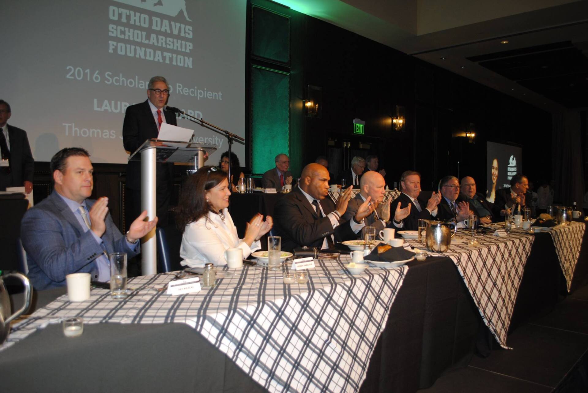 A group of people are sitting at a long table with a man at a podium giving a speech.