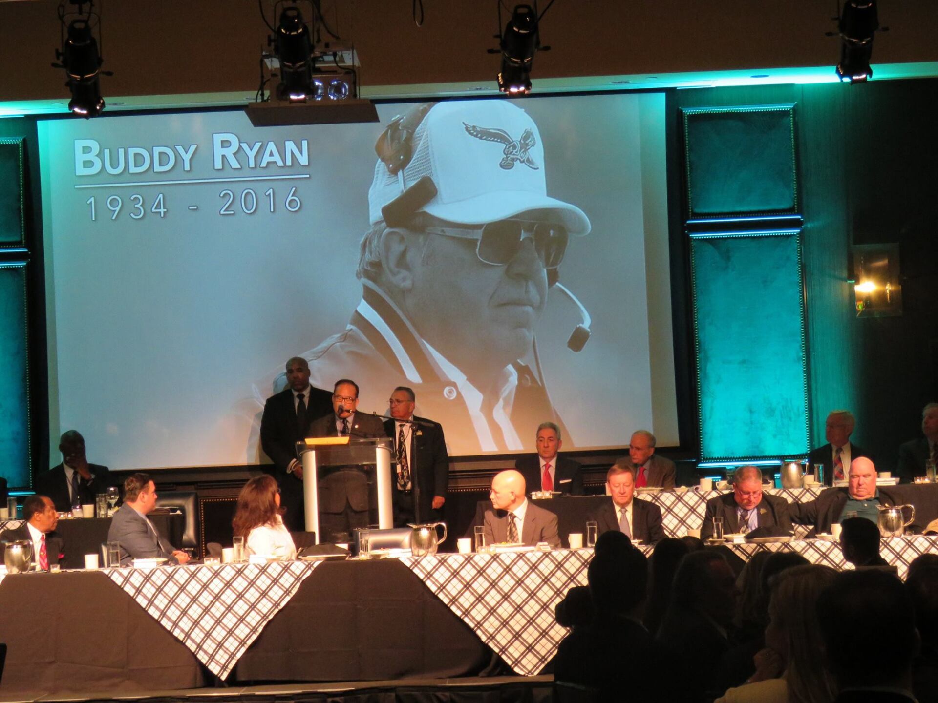 A group of people sitting at tables in front of a screen that says buddy ryan
