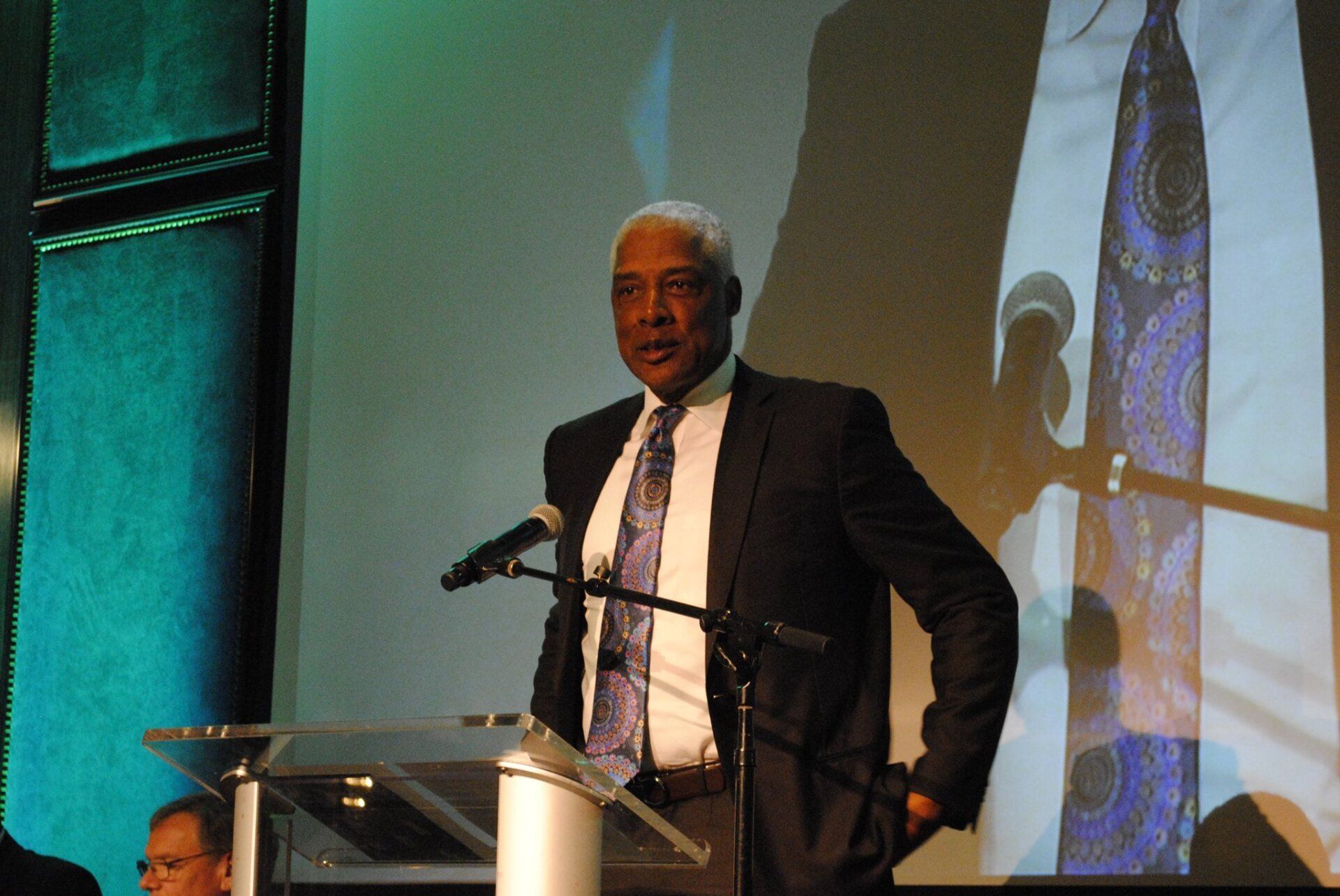 A man in a suit and tie stands at a podium speaking into a microphone