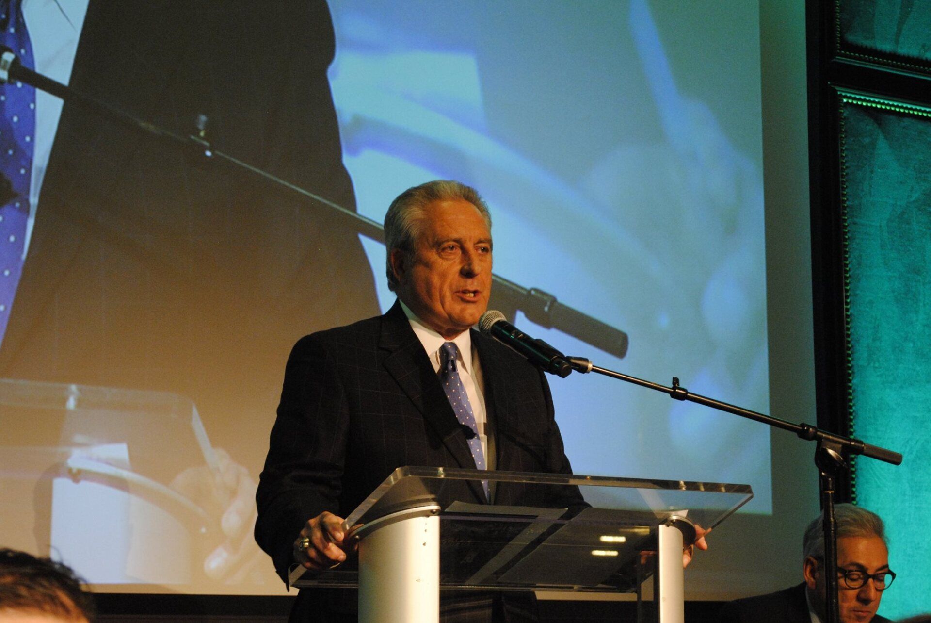 A man in a suit and tie is standing at a podium speaking into a microphone