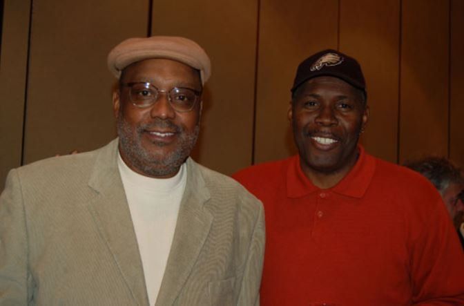 Two men are posing for a picture and one is wearing a hat