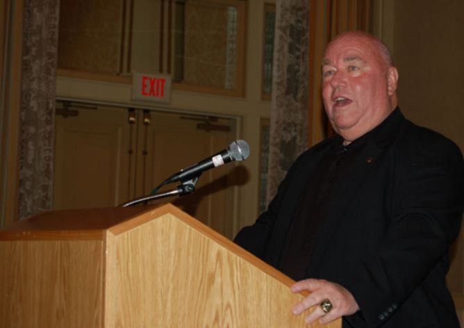 A man stands at a podium with a microphone in front of an exit sign