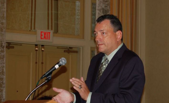 A man in a suit is speaking into a microphone in front of an exit sign