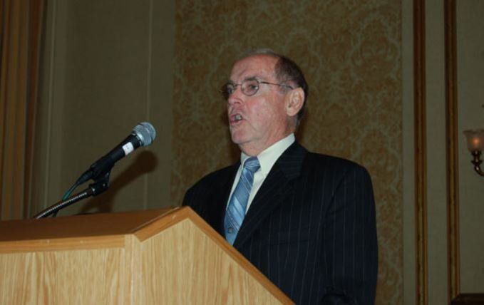 A man in a suit and tie stands at a podium speaking into a microphone
