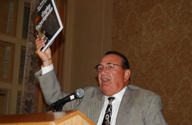 A man in a suit and tie is holding up a book while speaking into a microphone