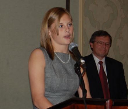 A woman is speaking into a microphone while a man sits behind her