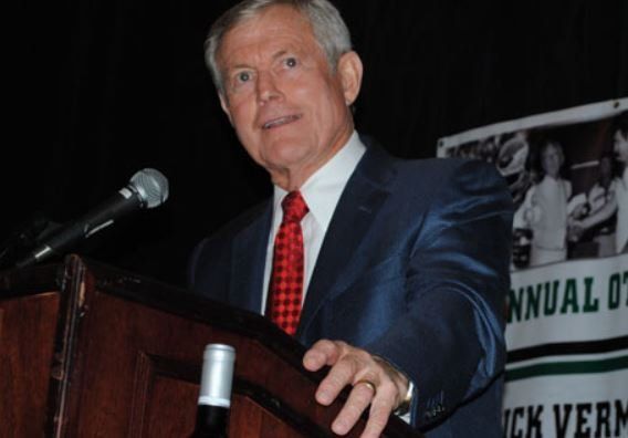 A man in a suit and tie stands at a podium holding a bottle of wine