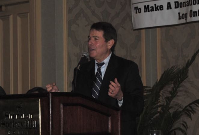 A man stands at a podium with a sign behind him that says to make a donation log on
