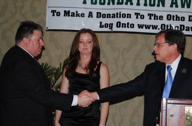 Two men shaking hands with a woman in front of a sign that says 
