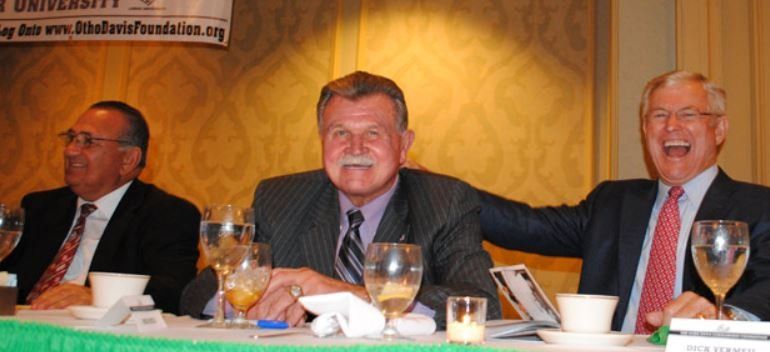 Three men are sitting at a table with wine glasses and a sign that says university