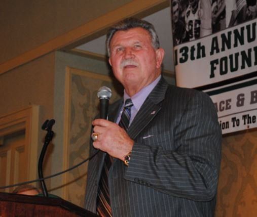 A man in a suit and tie is speaking into a microphone in front of a sign that says 13th annual found