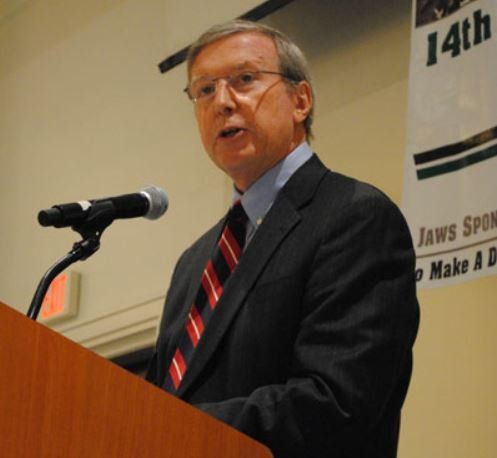 A man in a suit and tie stands at a podium speaking into a microphone