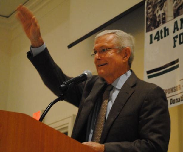 A man in a suit stands at a podium with his hand in the air