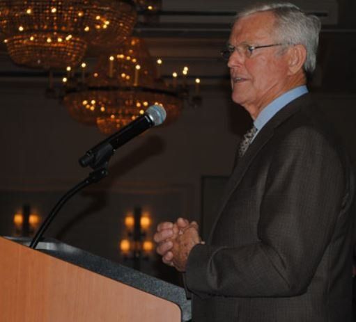 A man in a suit stands at a podium speaking into a microphone