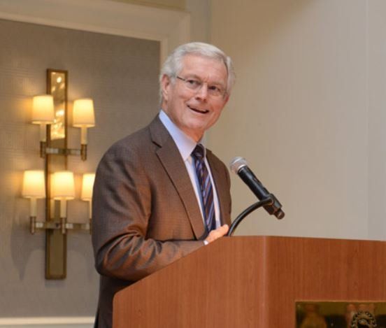 A man in a suit and tie stands at a podium speaking into a microphone