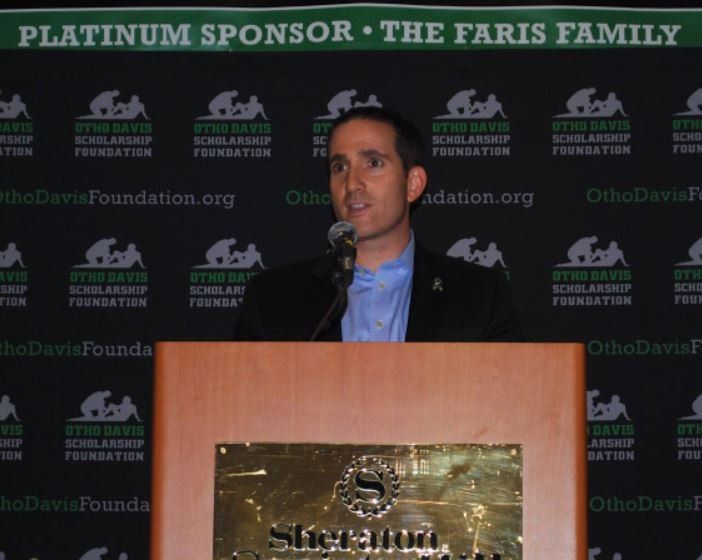 A man stands at a podium in front of a sign that says platinum sponsor the faris family