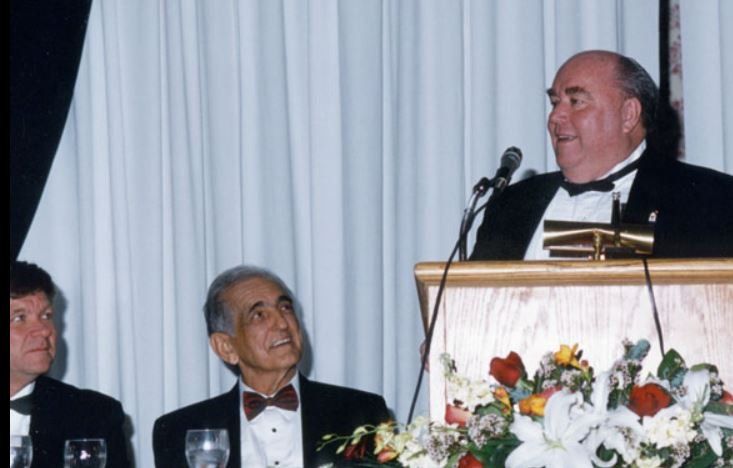 A man in a tuxedo is giving a speech at a podium