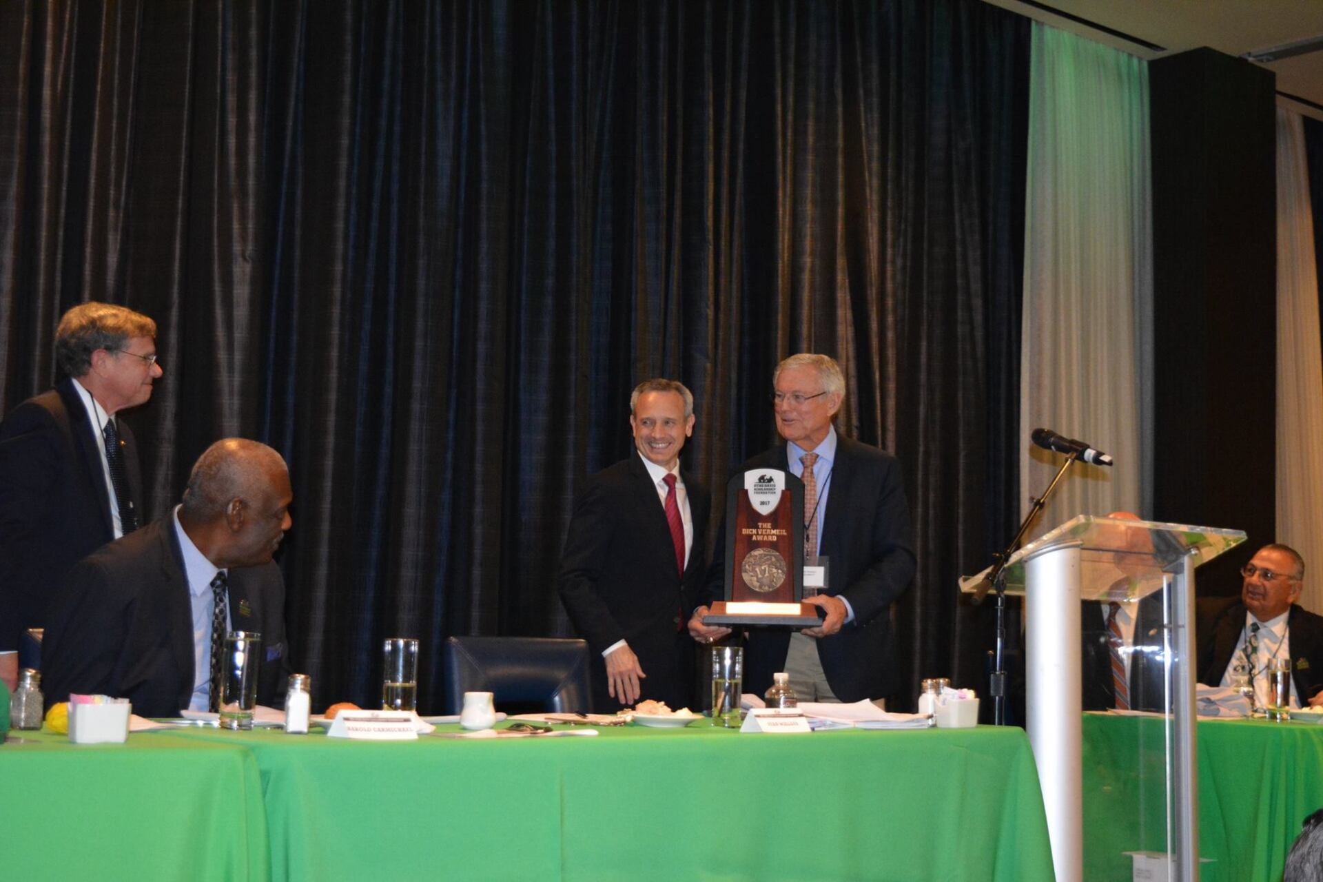 A man is holding a trophy in front of a group of men sitting at a table.