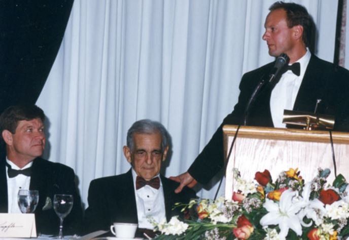 A man in a tuxedo stands at a podium giving a speech