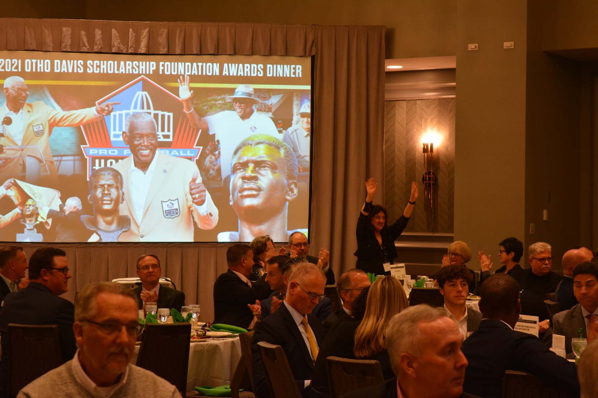 A group of people are sitting at tables in front of a large screen.