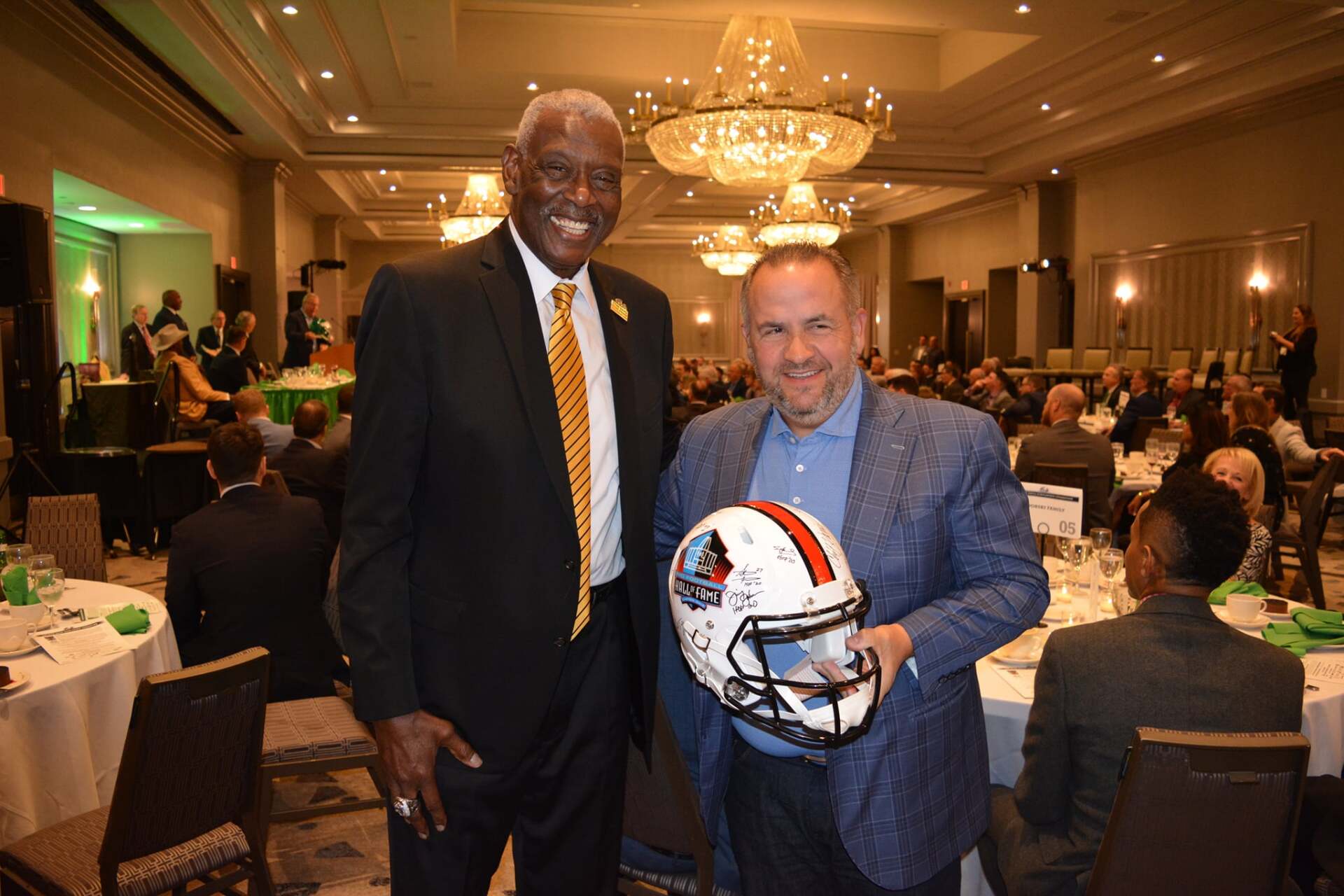 Two men are standing next to each other in a room holding a football helmet.