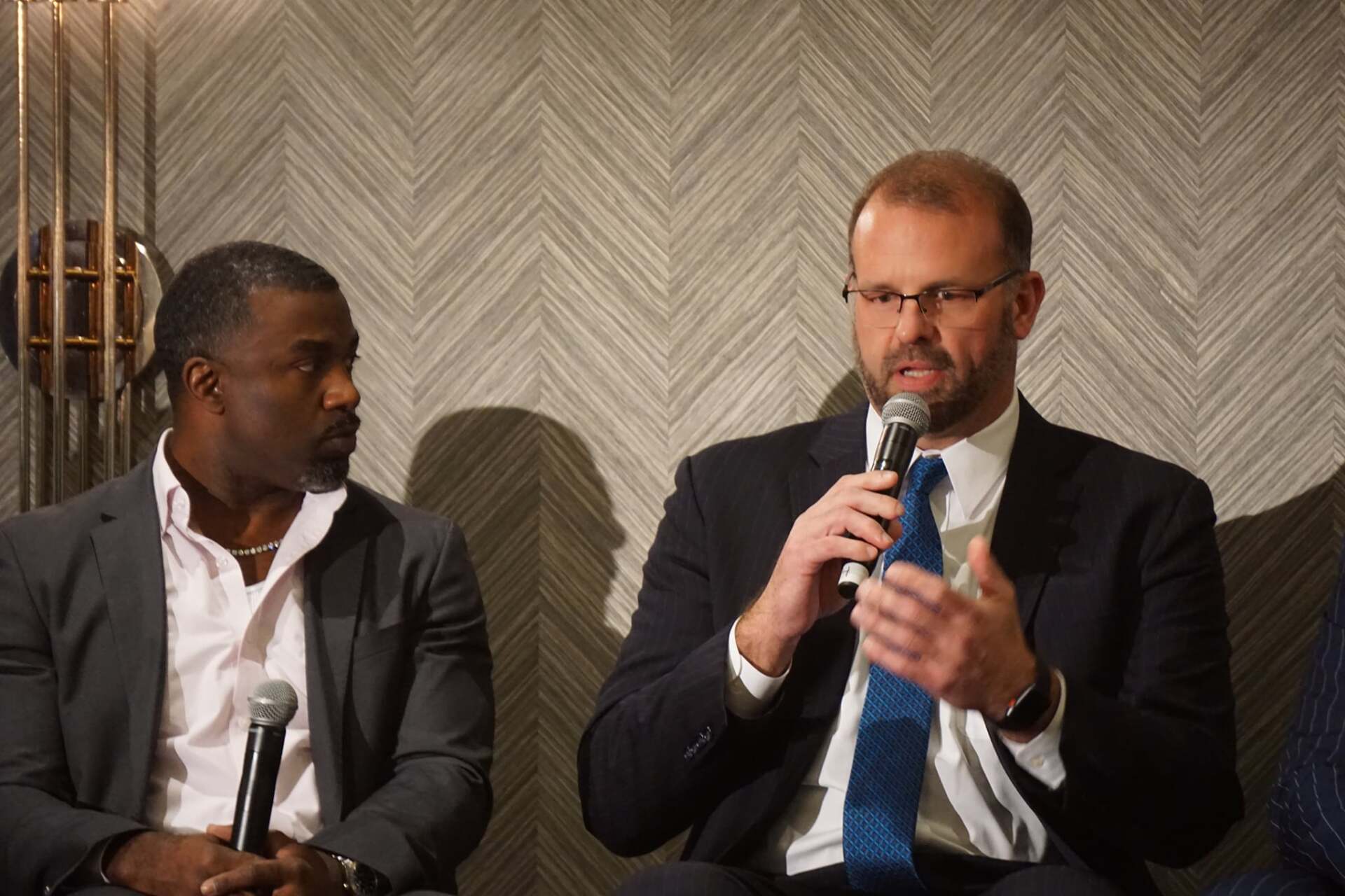 Two men in suits are sitting next to each other talking into microphones.