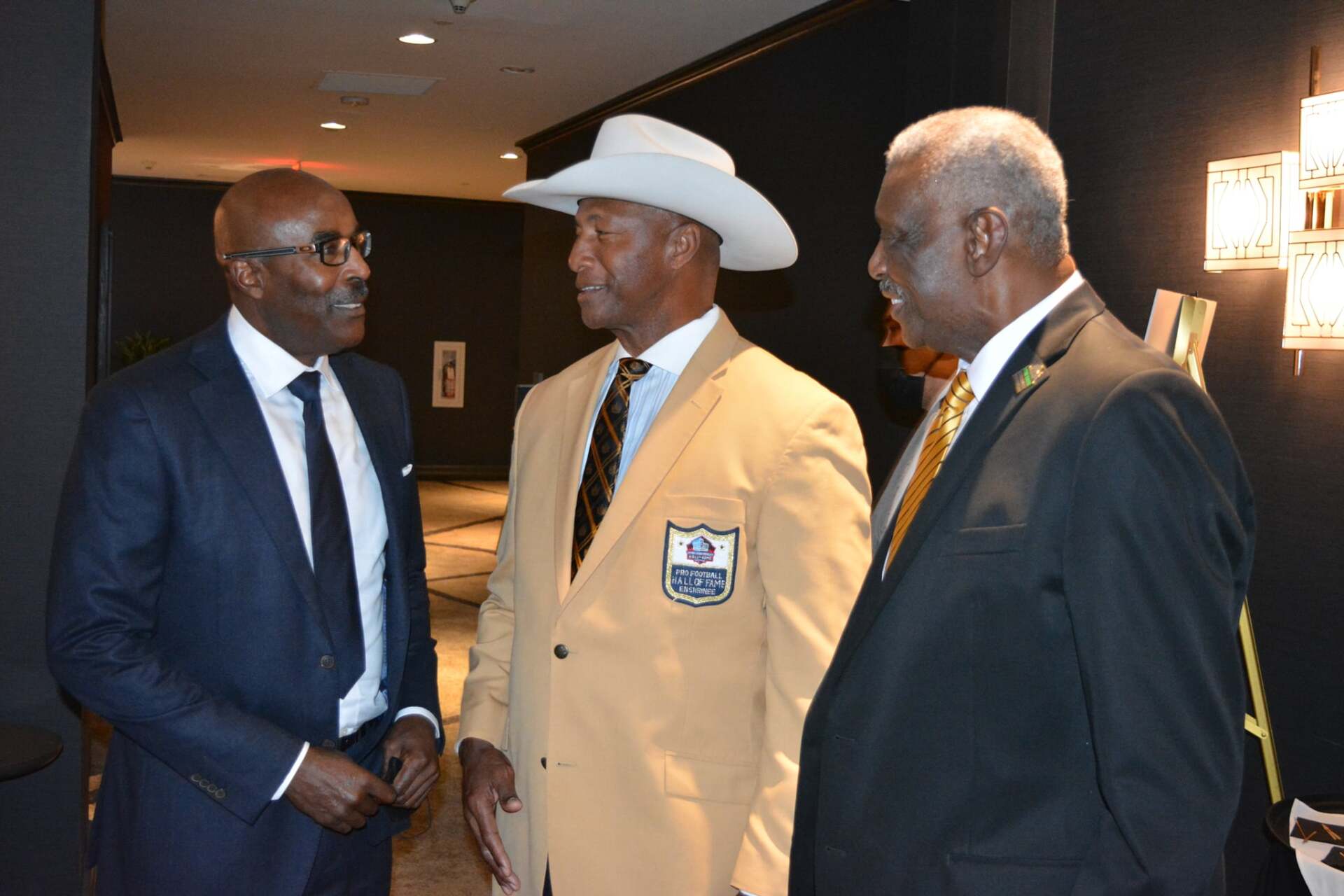 Three men in suits and ties are standing next to each other and talking . one of the men is wearing a cowboy hat.
