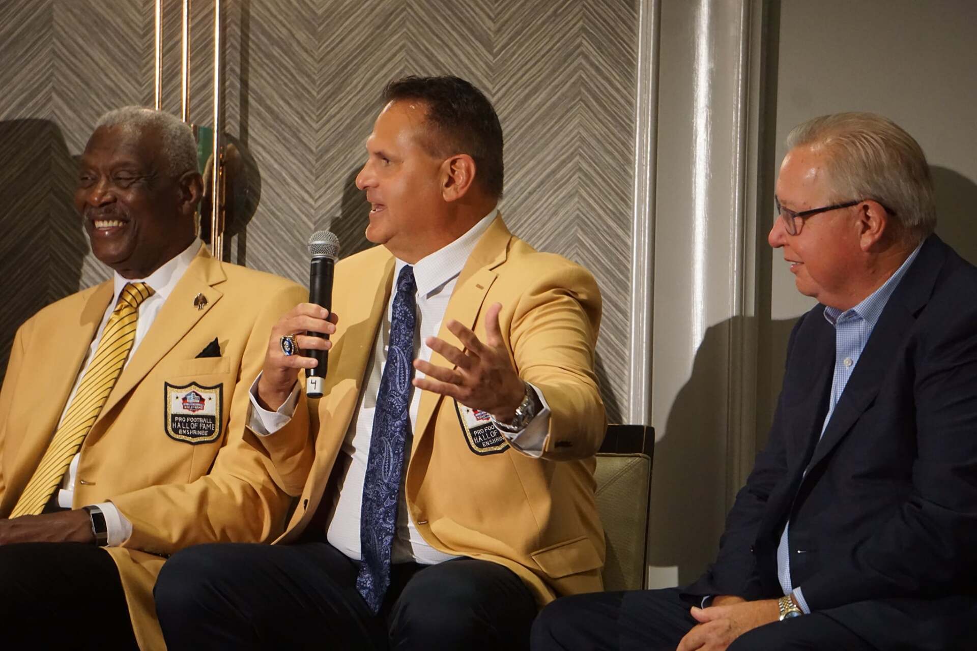 Three men in suits and jackets are sitting in a room and one of them is holding a microphone.