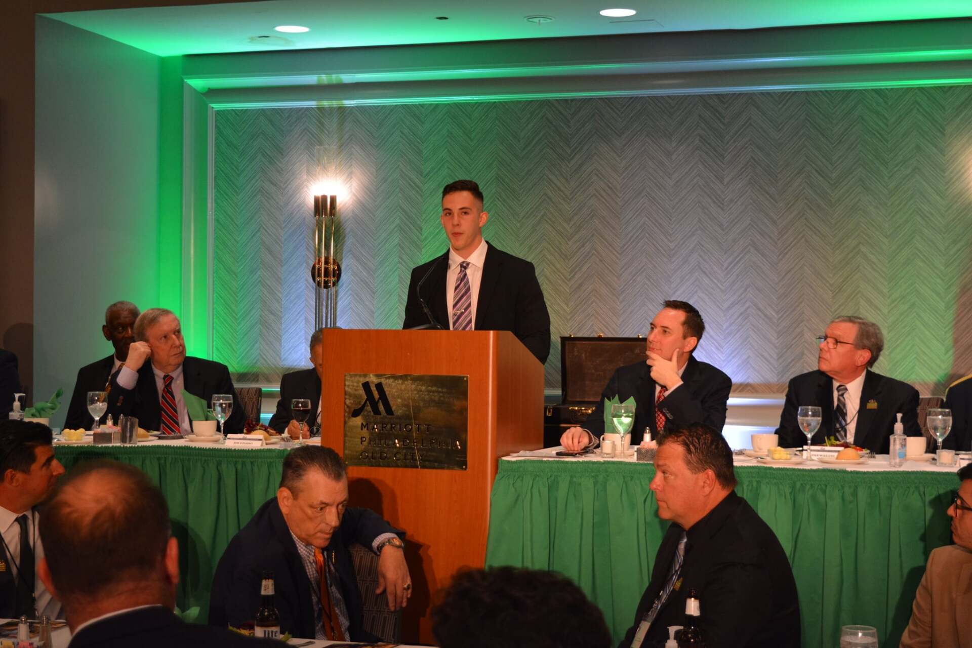 A man is standing at a podium giving a speech to a group of people sitting at tables.