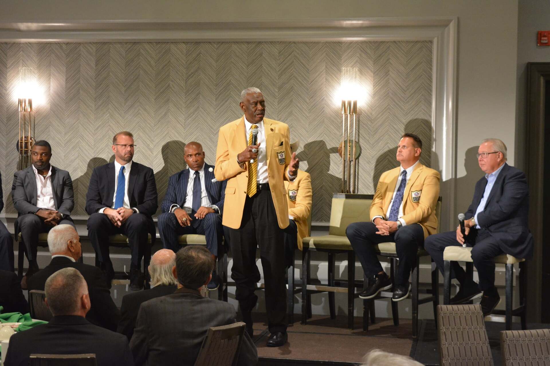 A man is standing on a stage talking into a microphone while a group of men sit in chairs.