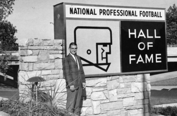 A man in a suit stands in front of a national professional football hall of fame sign