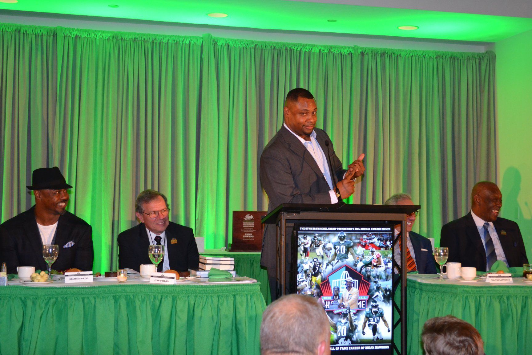 A man is standing at a podium giving a speech to a group of men sitting at tables.