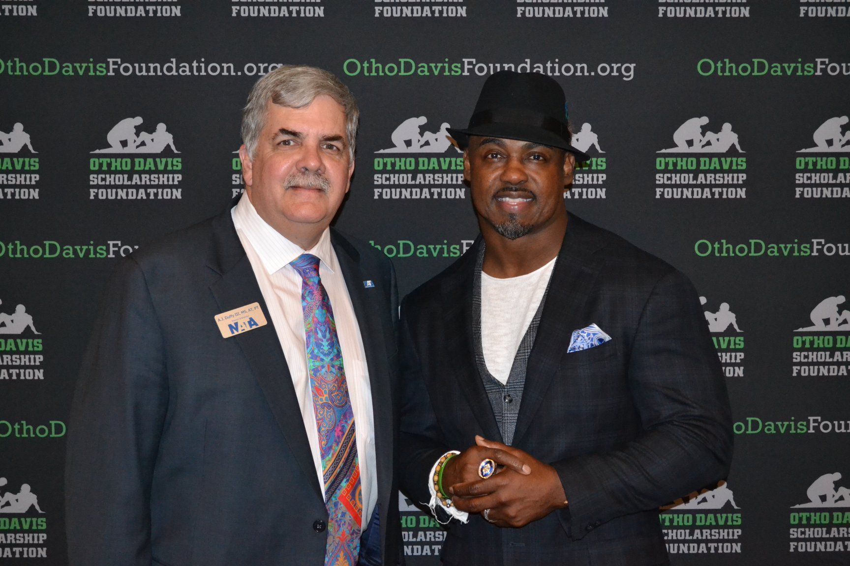 A man in a suit and tie is standing next to another man in a hat.