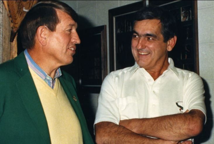 A man in a green jacket is talking to another man in a white shirt