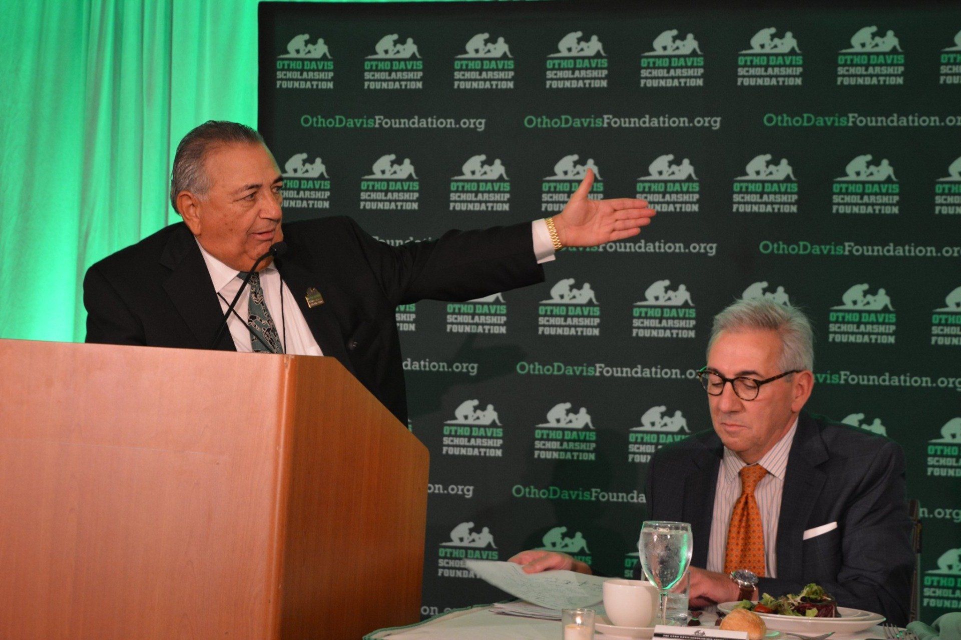 Two men are sitting at a table and one is giving a speech at a podium.